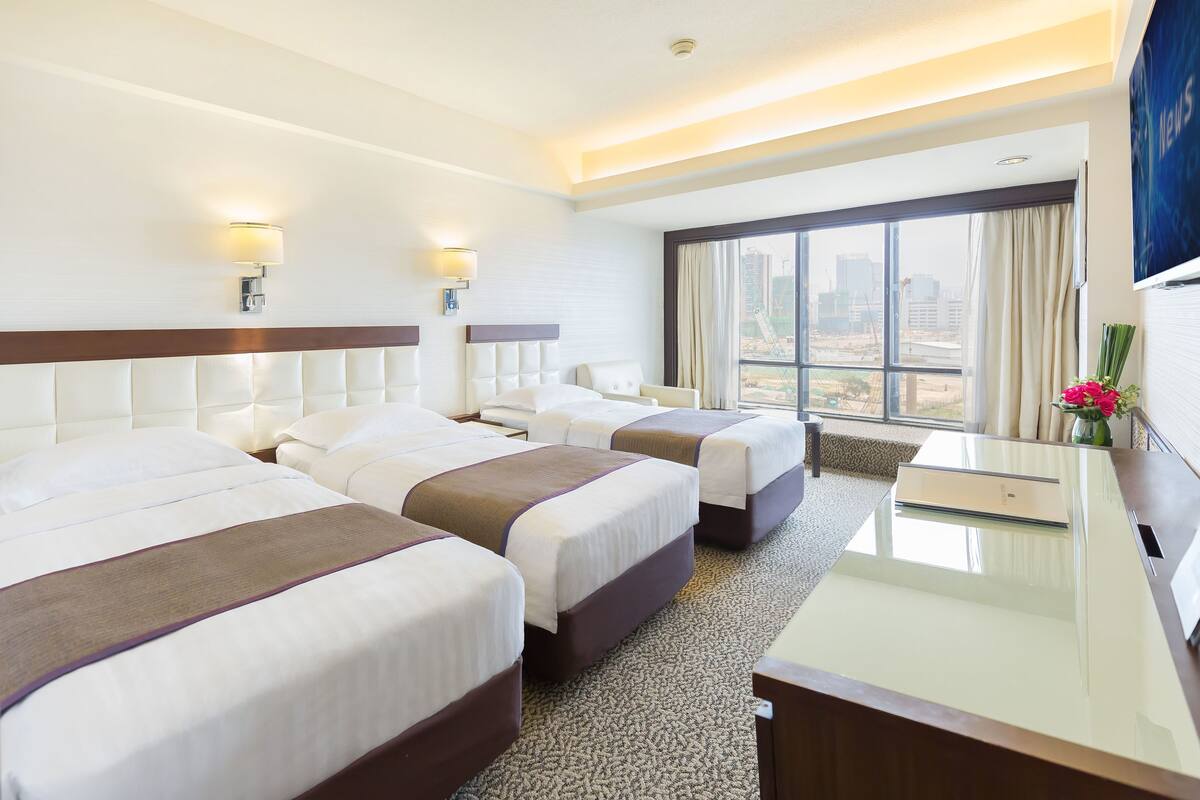A hotel room with three beds, a desk, and a large window with a city view.
