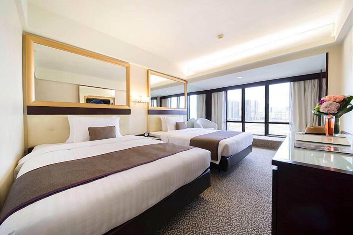 A hotel room with two beds, a desk, a large mirror, and a view of the city.