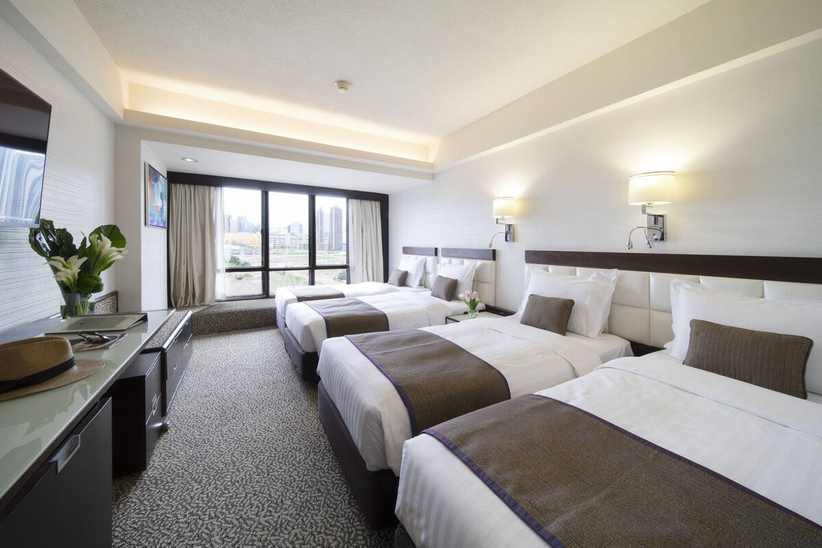 A hotel room with four beds, a desk, a large window with a city view, and a balcony.