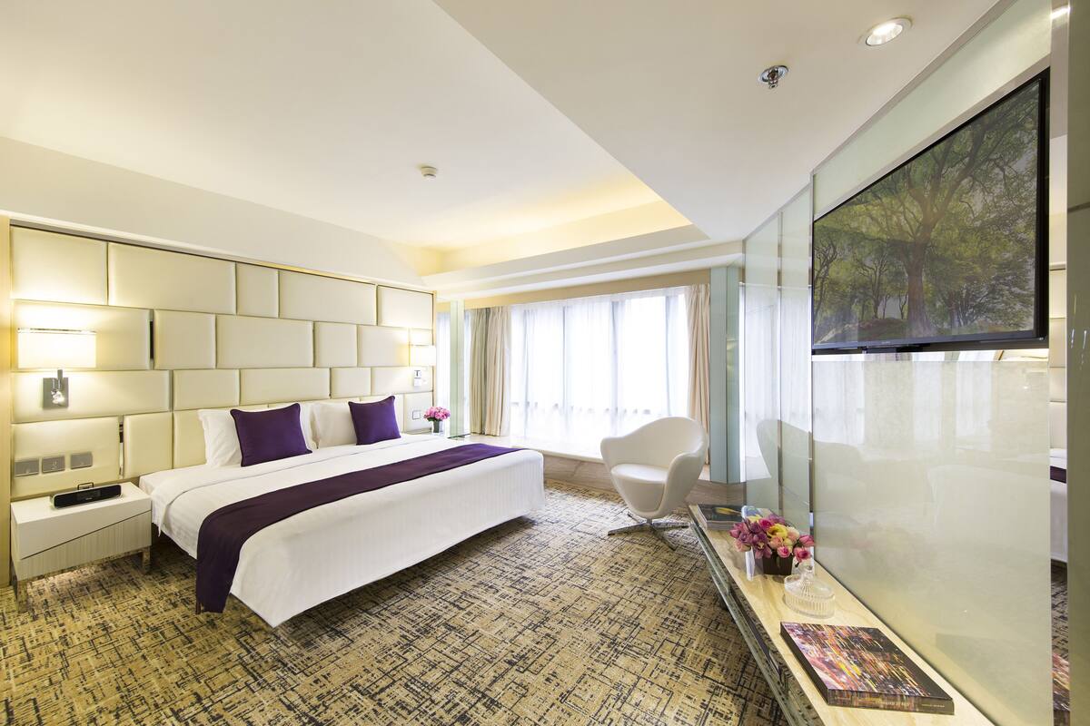 A modern hotel room with a large bed, a flat-screen TV, a desk with a chair, and a large window with curtains.