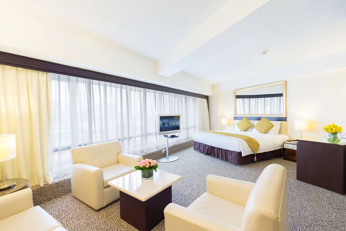 A hotel room with a bed, two white armchairs, a small table with a vase of flowers, a TV, and a desk with a lamp.