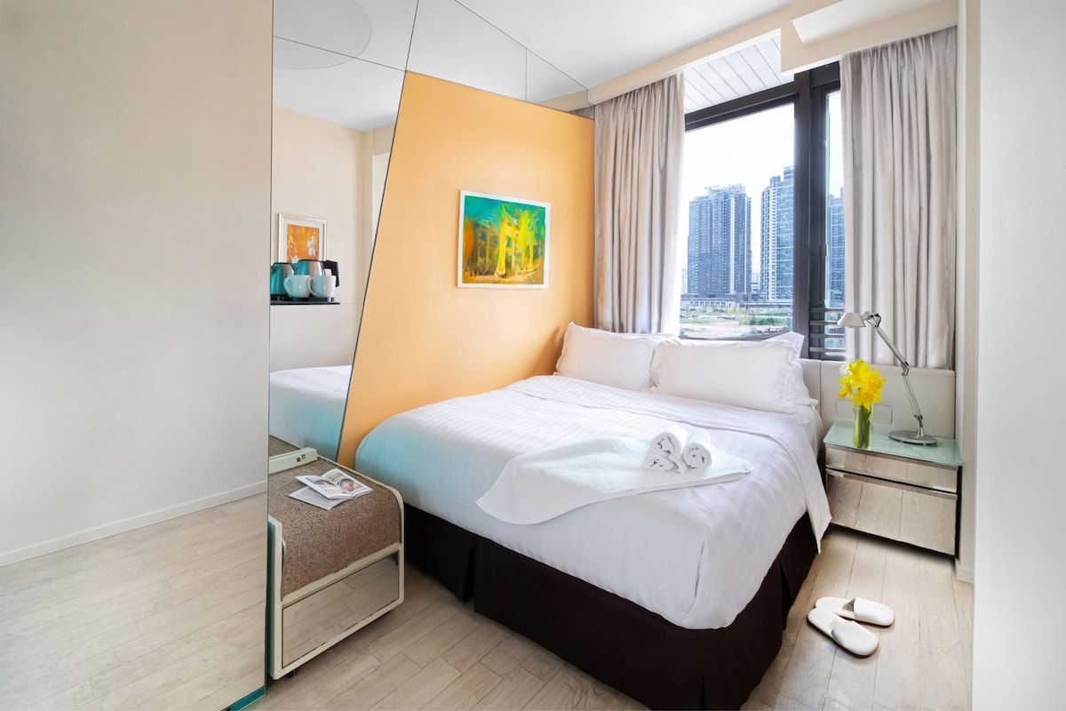 A hotel room with a bed, a bedside table with a lamp and flowers, a window with a city view, and a painting on the wall.