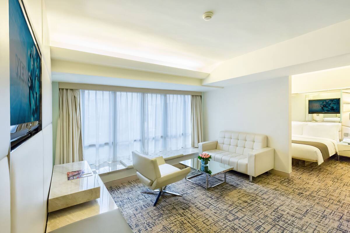 A hotel room with a sofa, a coffee table, a TV, a bed, and a large window with curtains.