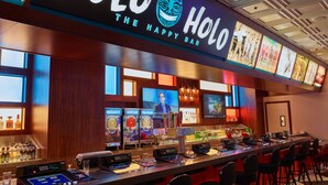 Un espace bar doté d’un écran numérique affichant un barman, d’un long comptoir avec des tabourets de bar, et d’une enseigne au-dessus indiquant « Holo, le bar où il fait bon vivre ».