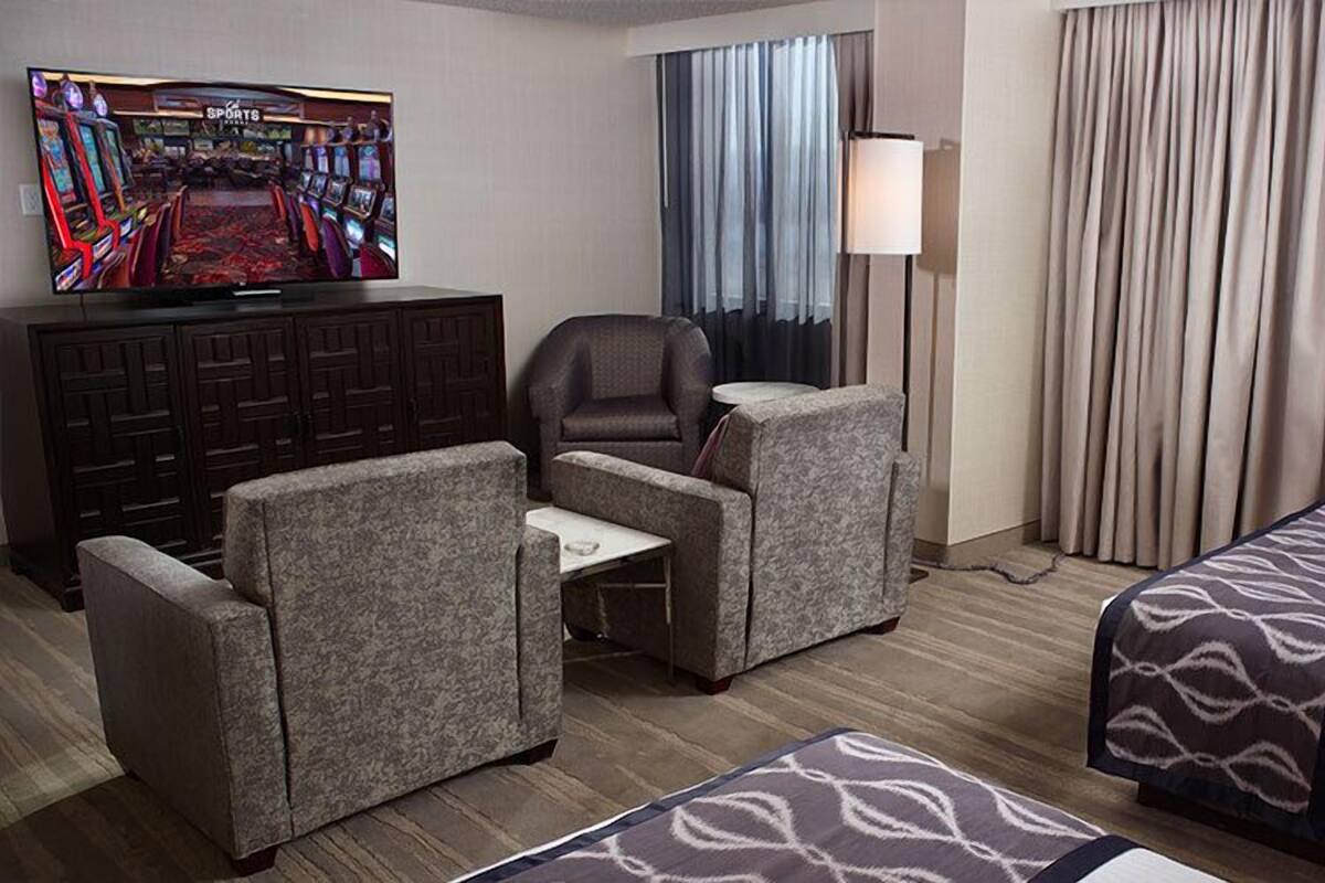 Ein Hotelzimmer mit Fernseher, der einen Sportkanal zeigt, einem Schreibtisch mit Stuhl, zwei Sofas, einem Bett mit gemusterten Bettwäsche, einer Lampe und Vorhängen.