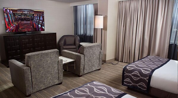 Una habitación de hotel con televisión mostrando un canal deportivo, un escritorio con silla, dos sofás, una cama con ropa de cama estampada, una lámpara y cortinas.