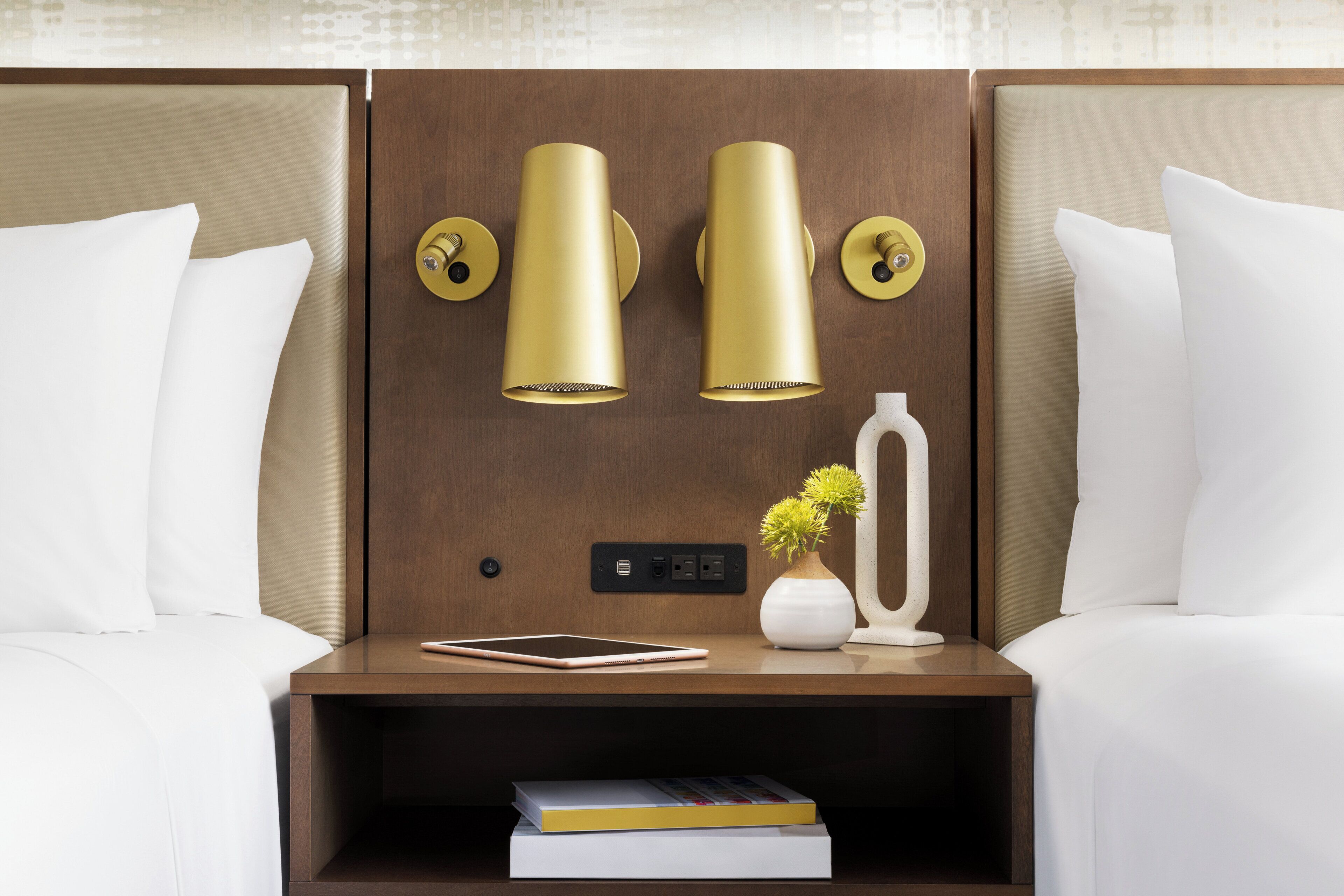 A hotel room with a wooden headboard, two bedside lamps, a bedside table with a book and a vase with flowers, and a power outlet.