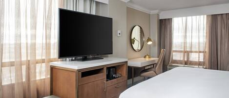 Et hotelværelse med en seng, et fjernsyn på en trækommode, et skrivebord med lampe og et stort vindue med gardiner.