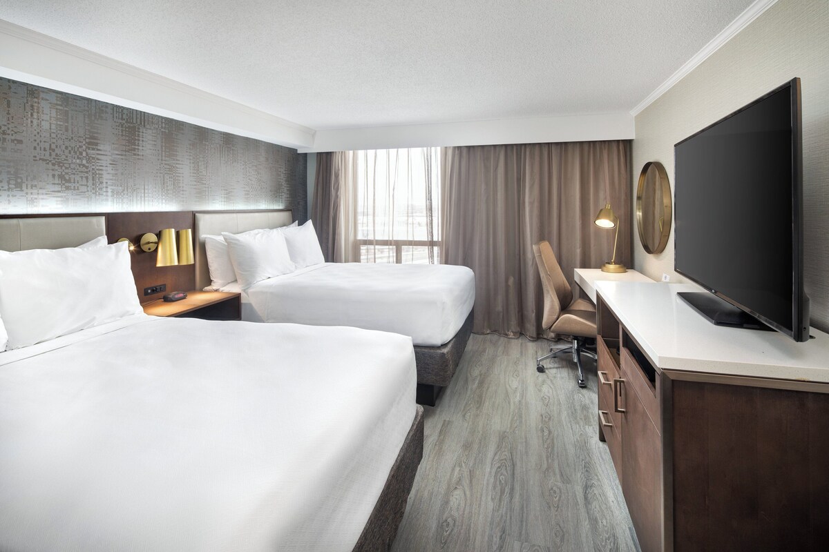 Habitación de hotel con dos camas, un escritorio, un televisor y un ventanal con cortinas.