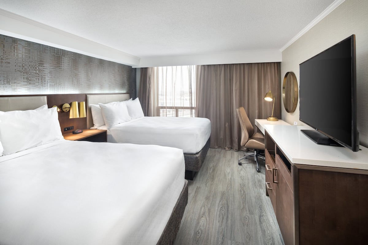 Ein Hotelzimmer mit zwei Betten, einem Schreibtisch, einem Fernseher und einem großen Fenster mit Vorhängen.