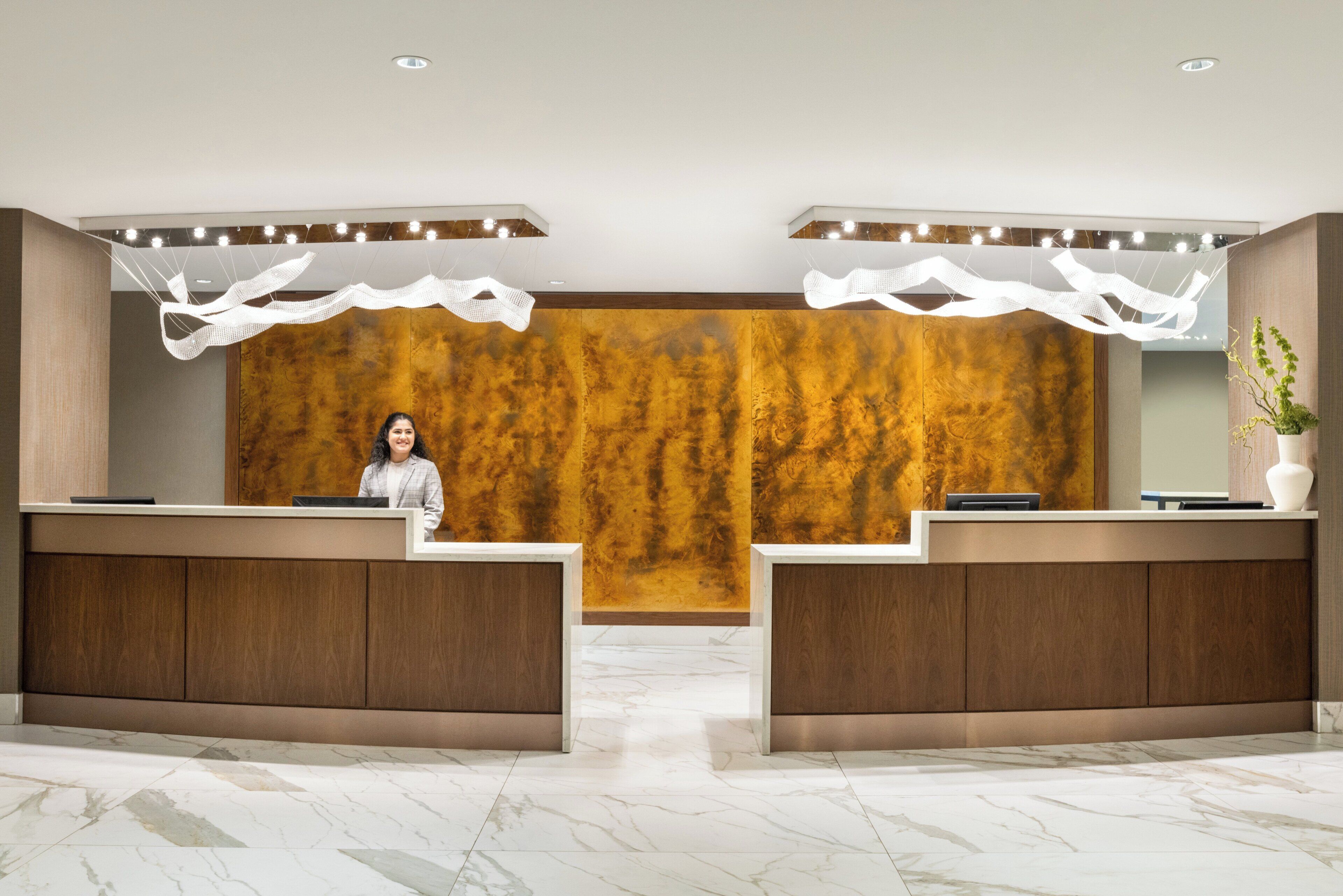 A reception desk with a woman standing behind it, a wooden wall with a textured pattern, and a plant on the right side.