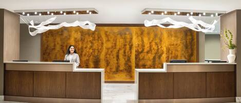 A reception desk with a woman standing behind it, a wooden wall with a textured pattern, and a plant on the right side.