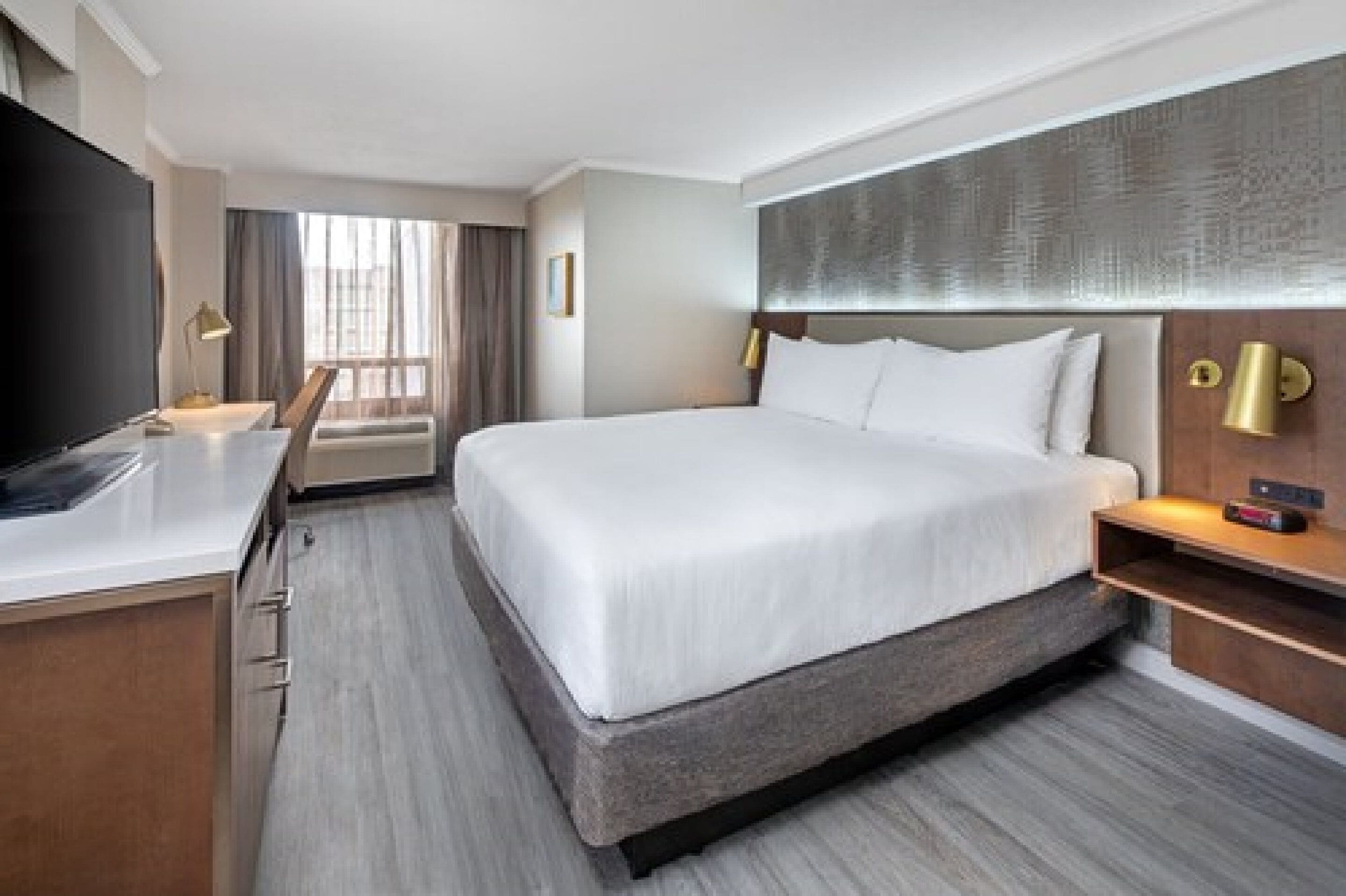 Une chambre d’hôtel avec un grand lit, un bureau doté d’un téléviseur à écran plat, une table de chevet avec une lampe, et une vue sur la ville par la fenêtre.