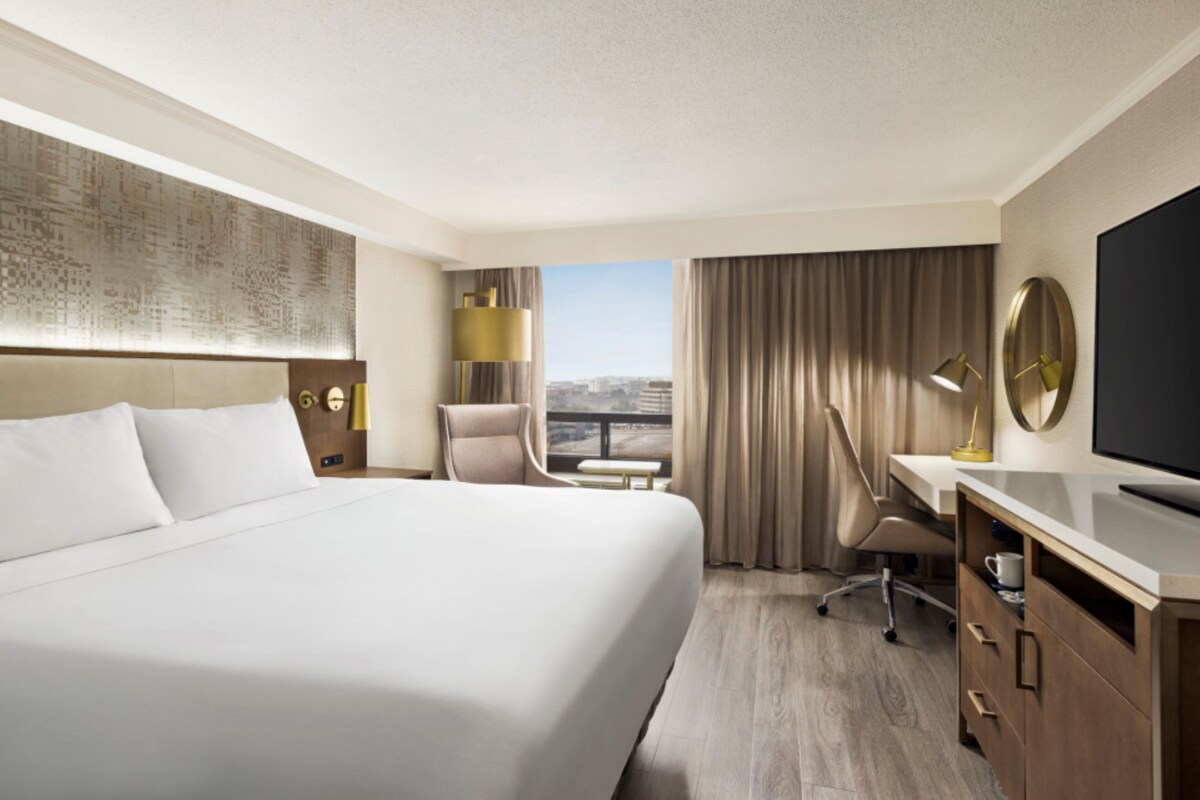 Habitación de hotel con una cama grande, un escritorio, una silla, un televisor y vistas a la ciudad.