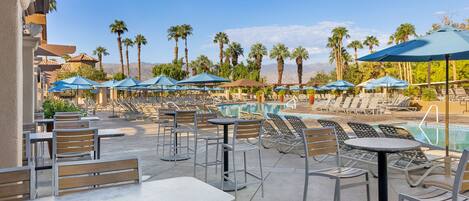 A poolside area with lounge chairs and umbrellas, surrounded by palm trees and mountains in the background.