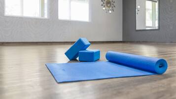 Un tapis de yoga, deux rouleaux de mousse et un bloc de yoga sur un plancher en bois, dans une pièce dotée de grandes fenêtres et d’un miroir décoratif.
