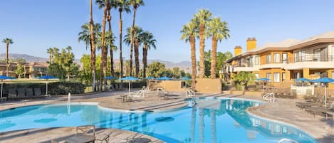 A swimming pool with lounge chairs and umbrellas, surrounded by palm trees and a building with balconies.