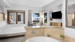 Een moderne hotelkamer met een groot bed, een badkamer met bad en een televisie, en uitzicht op palmbomen.