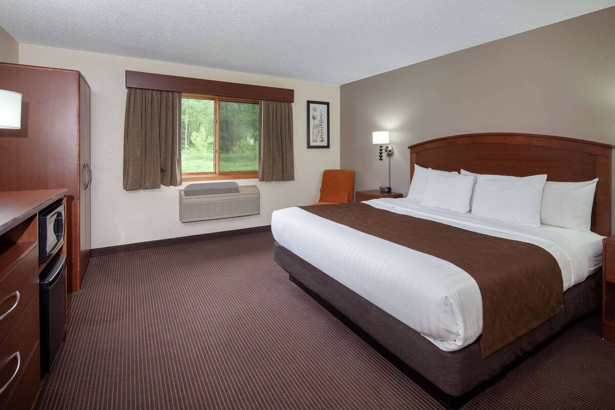 Ein Hotelzimmer mit einem großen Bett, einem Schreibtisch mit Stuhl, einem Schminktisch, einem Fenster mit Vorhängen und einer Wand-Klimaanlage.