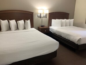 A hotel room with two beds, each with white bedding and a wooden headboard. There is a nightstand with a lamp between the beds.
