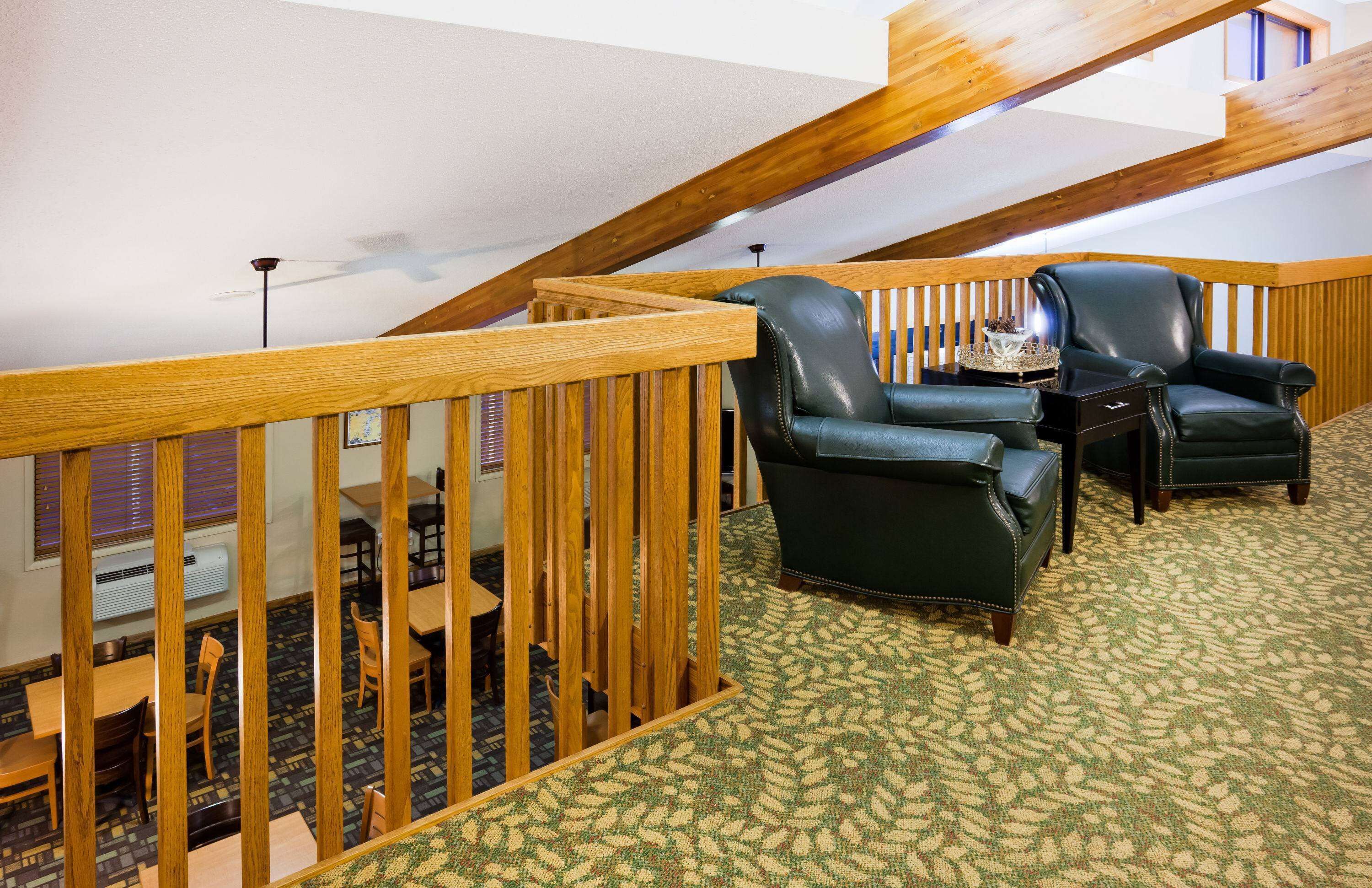 A high-ceilinged room with a wooden railing, two leather armchairs, a small table, and a patterned carpet.