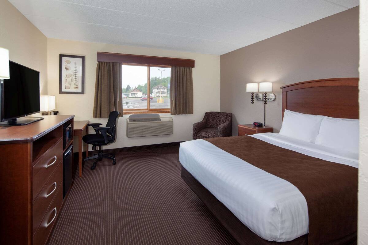 Ein Hotelzimmer mit einem großen Bett, einem Schreibtisch mit Fernseher, einem Sessel, einer Klimaanlage und einem Fenster mit Vorhängen.