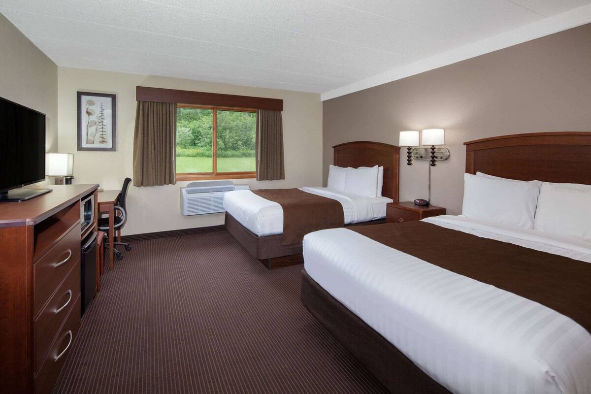 Ein Hotelzimmer mit zwei Betten, einem Schreibtisch mit Computer, einem Fernseher und einem Fenster mit Vorhängen.