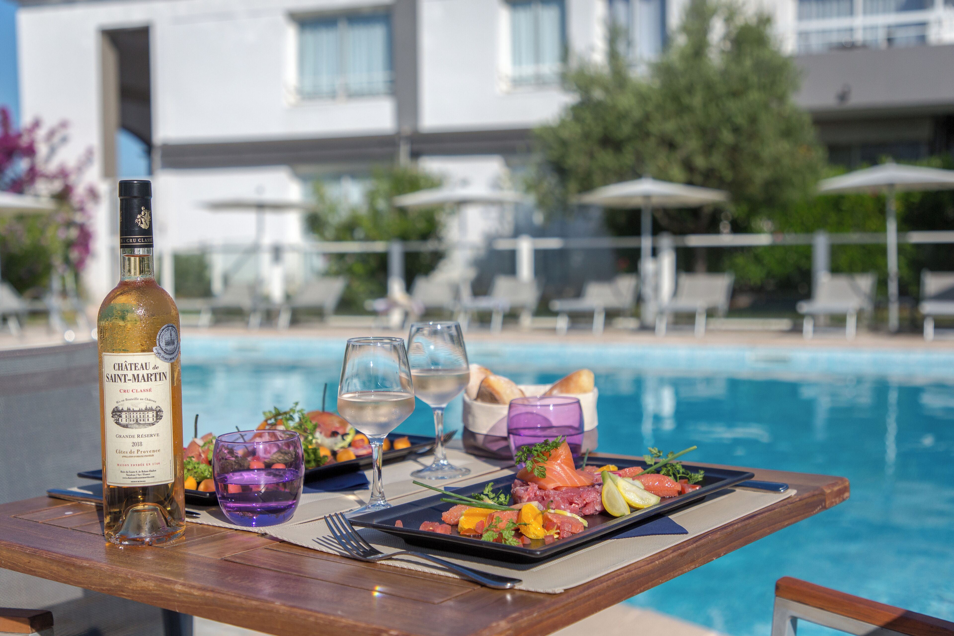Une table à manger avec une bouteille de vin du Château de Saint-Martin, deux verres de vin blanc, une assiette de nourriture et un panier de pain, le tout placé près d’une piscine.