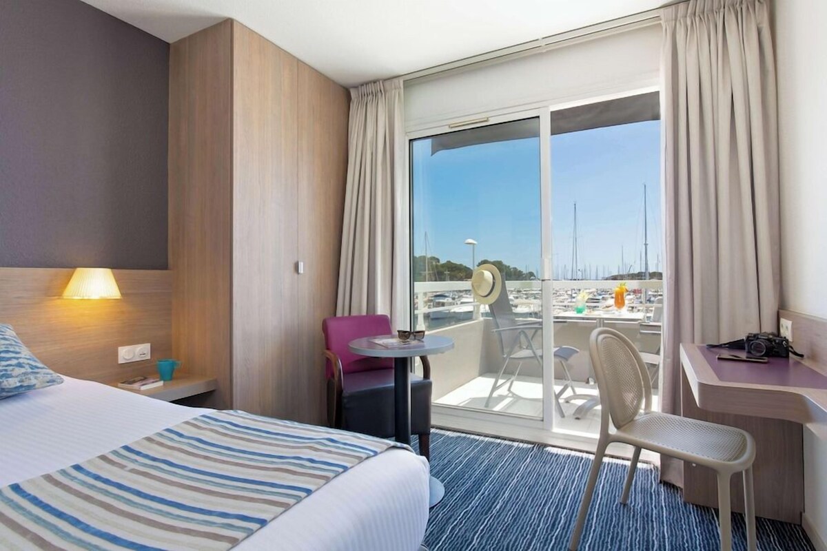 A hotel room with a bed, a desk, a chair, a small table, and a balcony with a view of the marina.