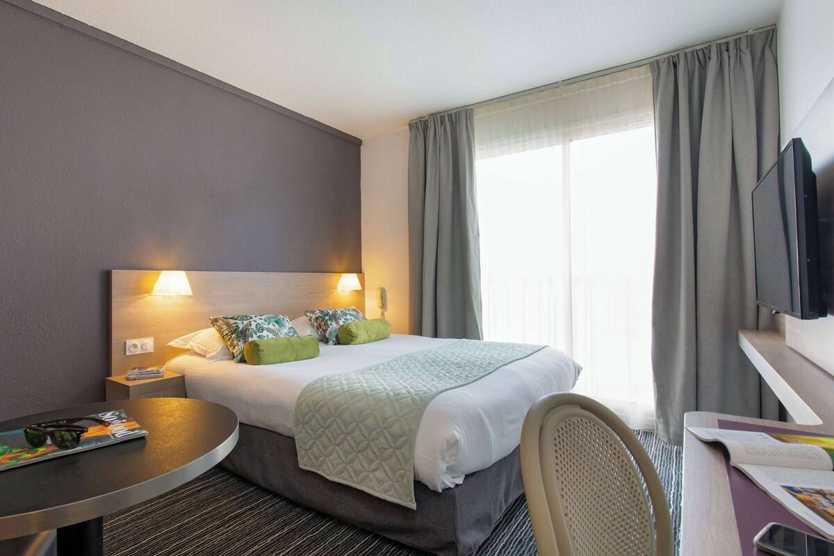 Een hotelkamer met een bed, nachtkastje, stoel, televisie en een raam met gordijnen.