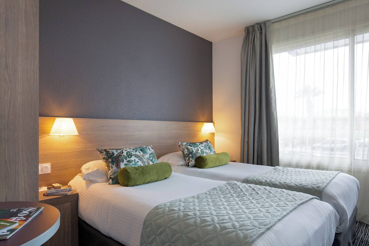 Een hotelkamer met twee bedden, een nachtkastje, een lamp en een raam met gordijnen.