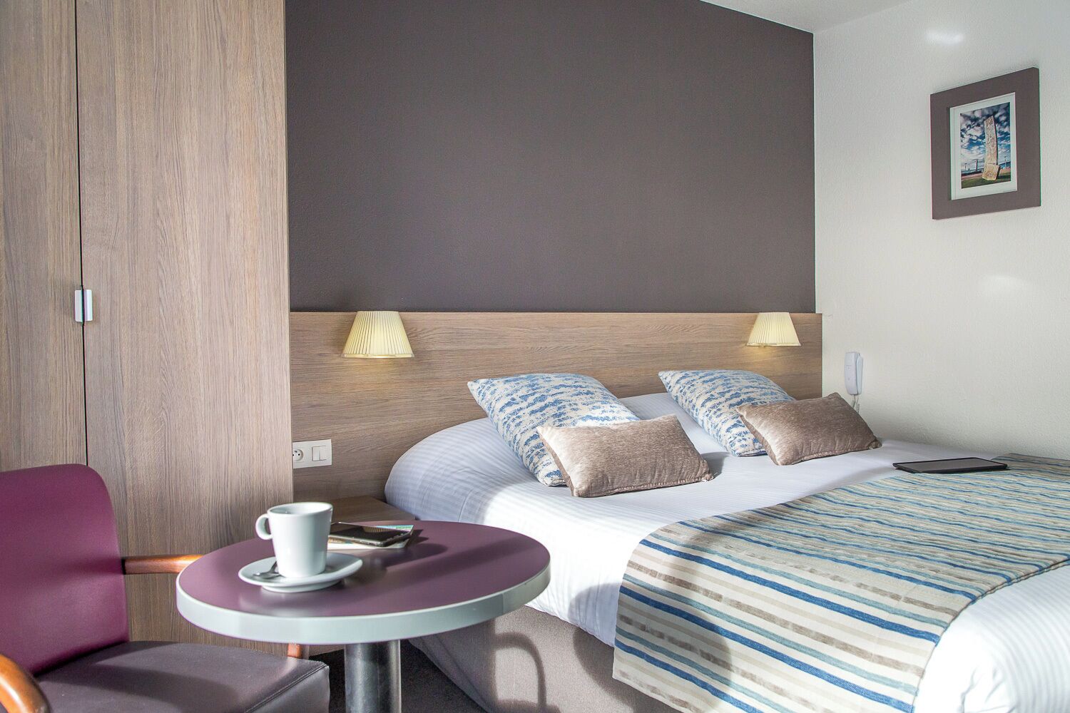Una habitación de hotel con una cama, lámparas de noche, una mesita con una taza y un plato, y una silla.