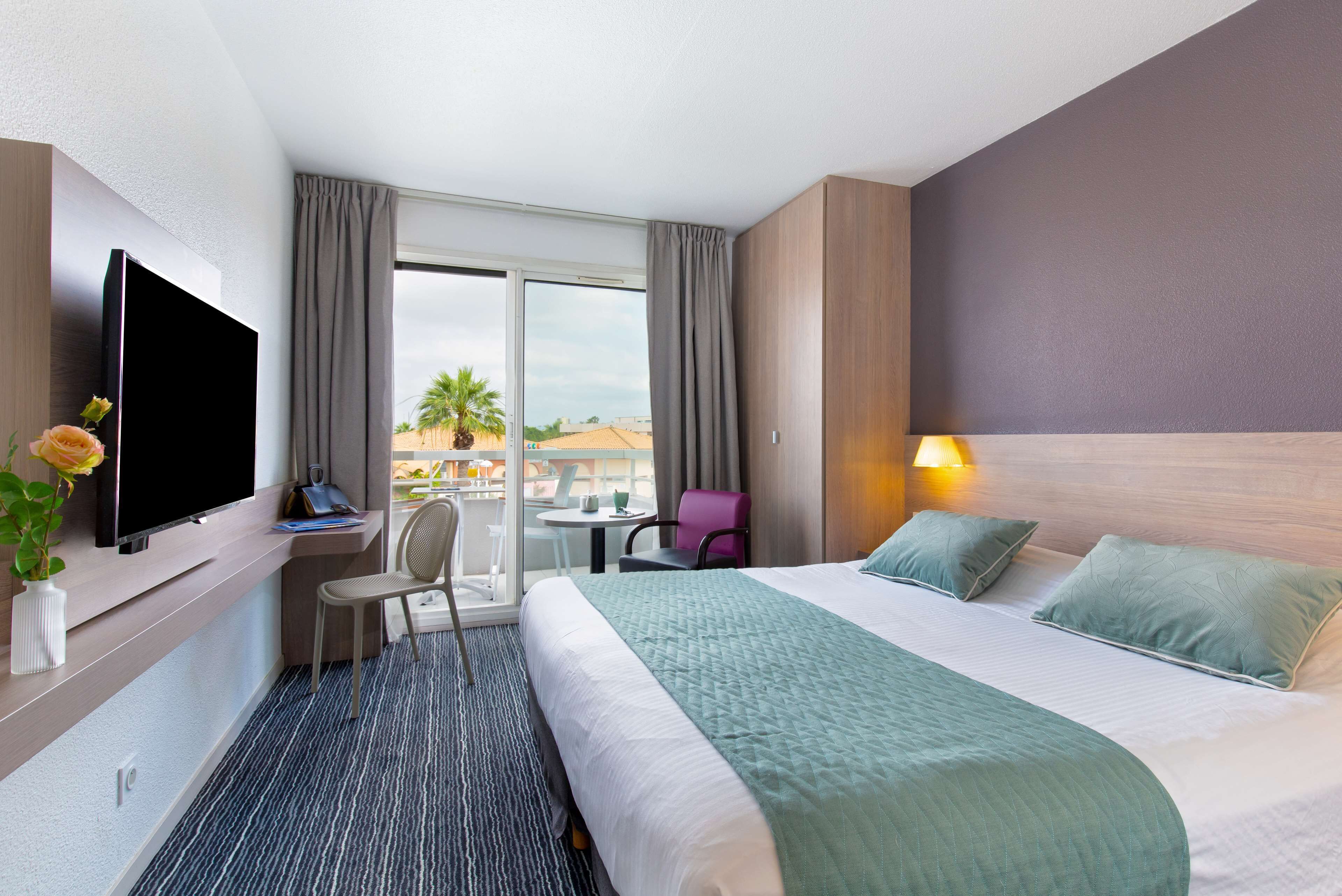 A hotel room with a bed, a desk with a computer, a TV, a chair, a small table, and a window with curtains.