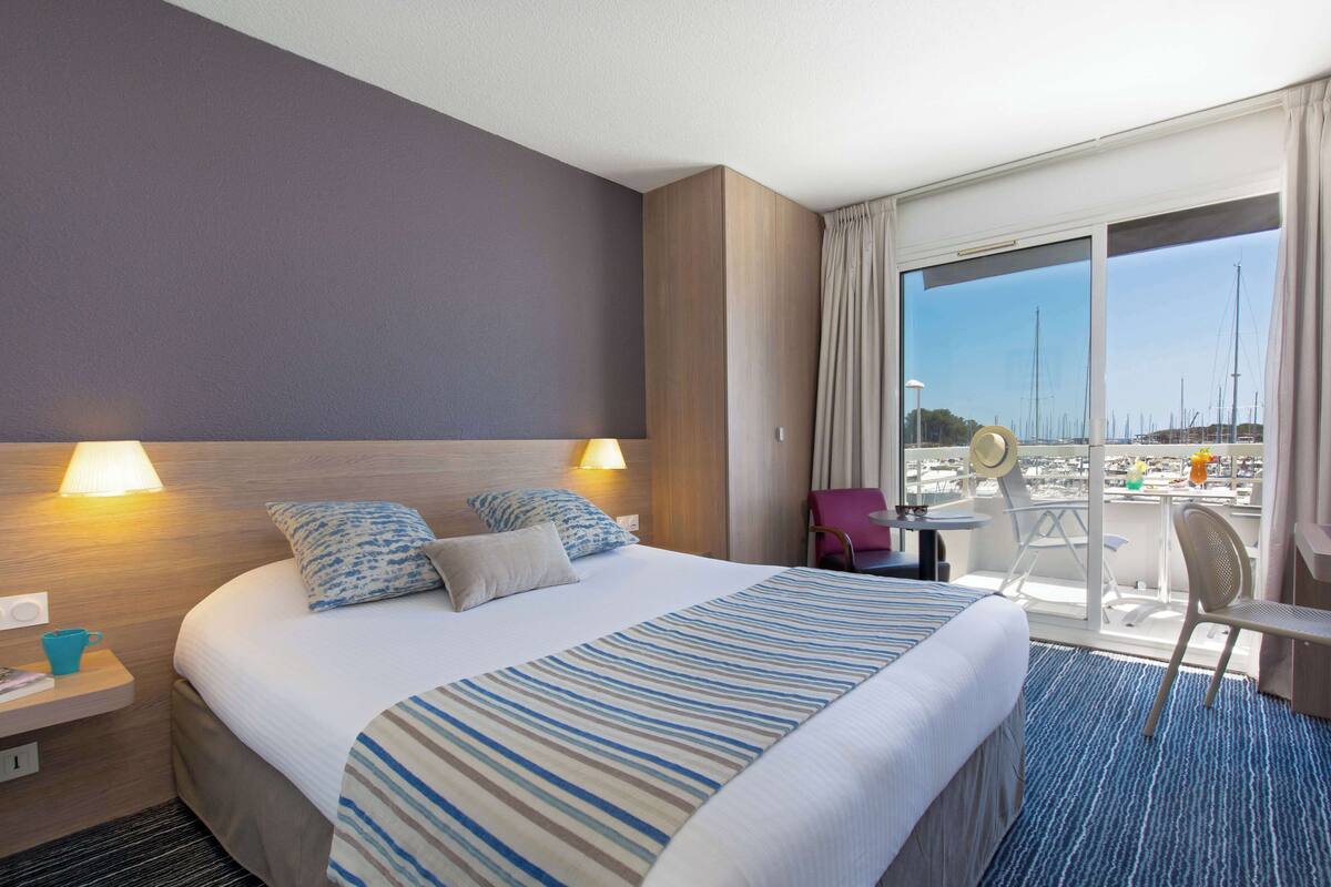 Een hotelkamer met een groot bed, twee bedlampen, uitzicht op de jachthaven en een balkon met een stoel en tafel.