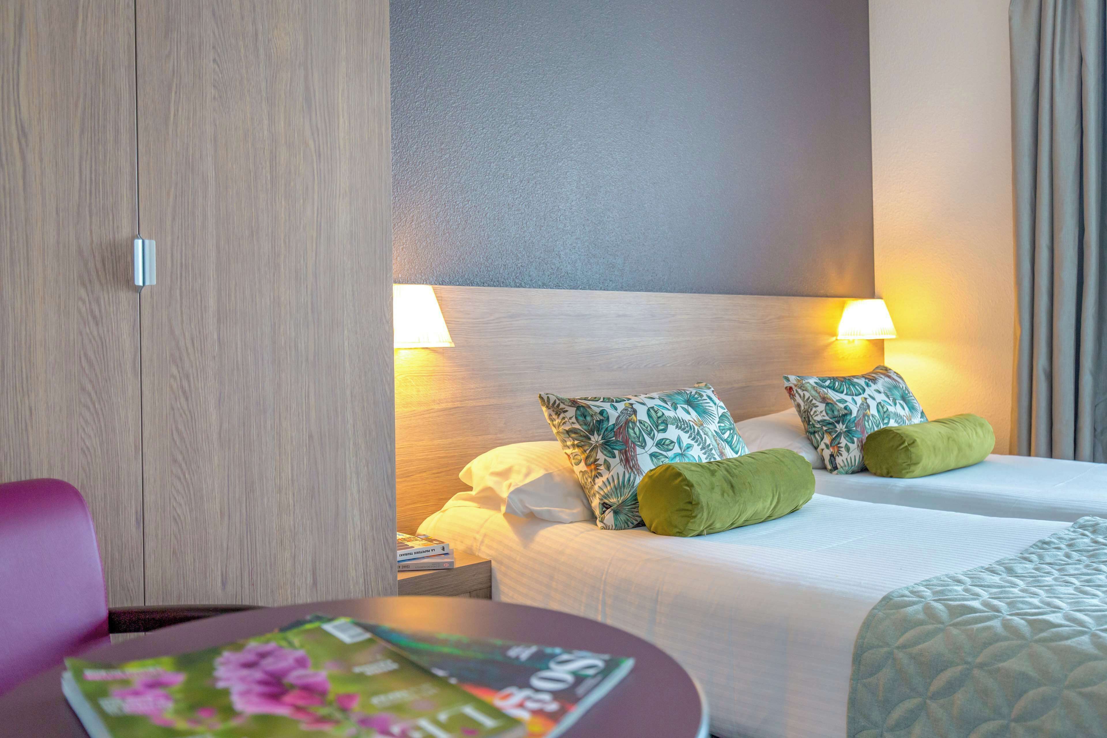 Une chambre d’hôtel avec un lit, deux oreillers, une table de chevet, un magazine et une chaise.