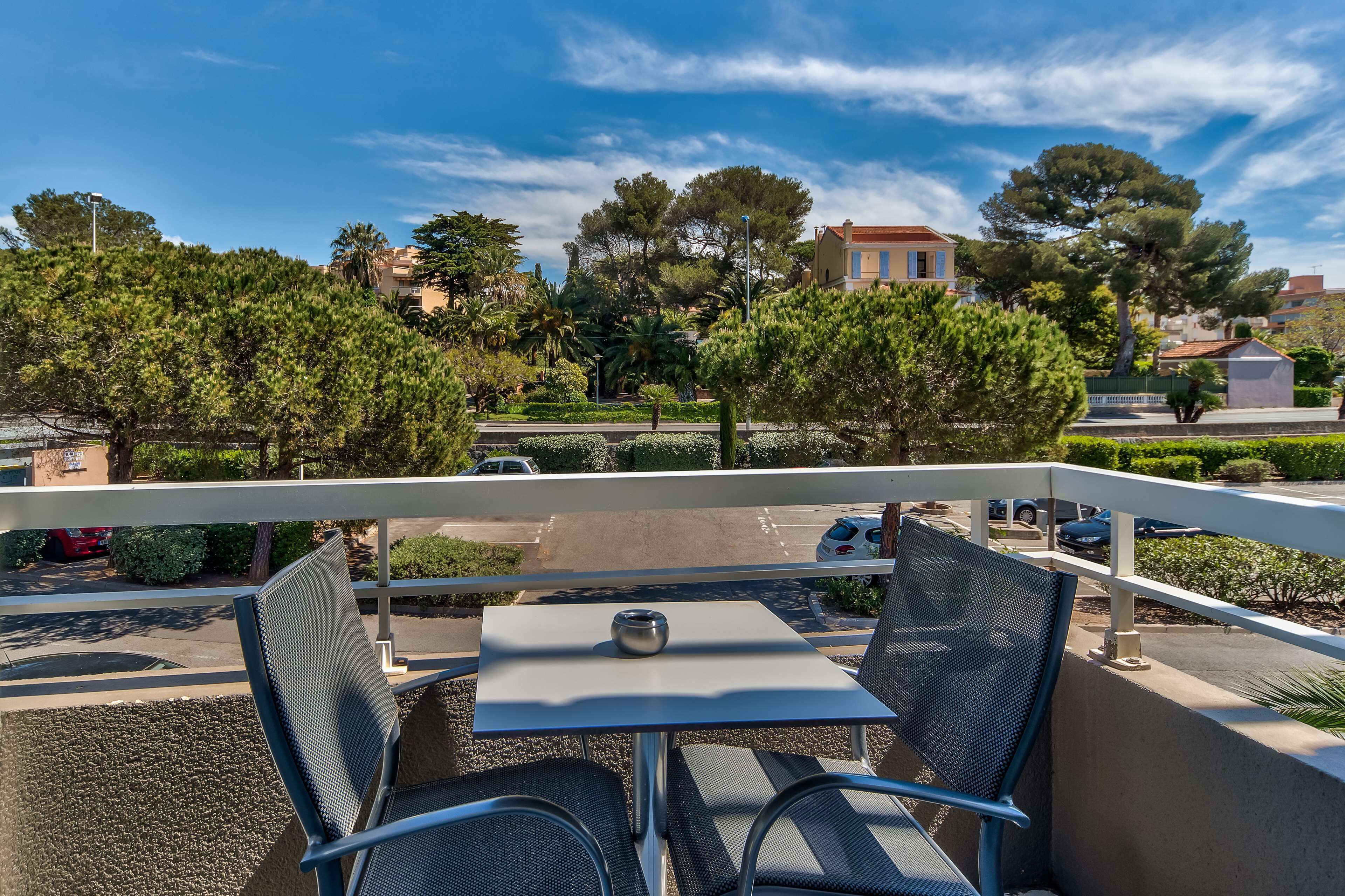 A balcony with two chairs and a table, overlooking a street with parked cars and trees.