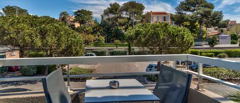 A balcony with two chairs and a table, overlooking a street with parked cars and trees.