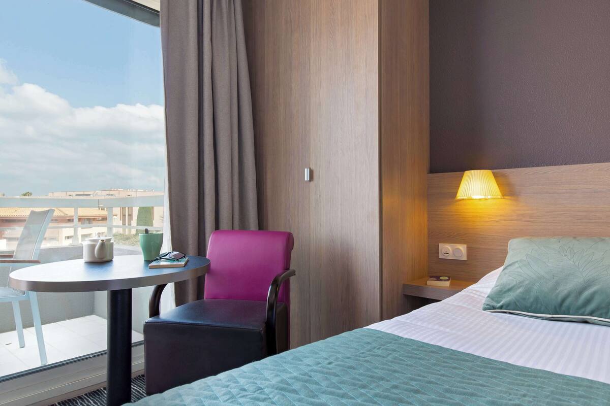 Een hotelkamer met een bed, een tafel met stoel, een lamp en uitzicht op de stad.