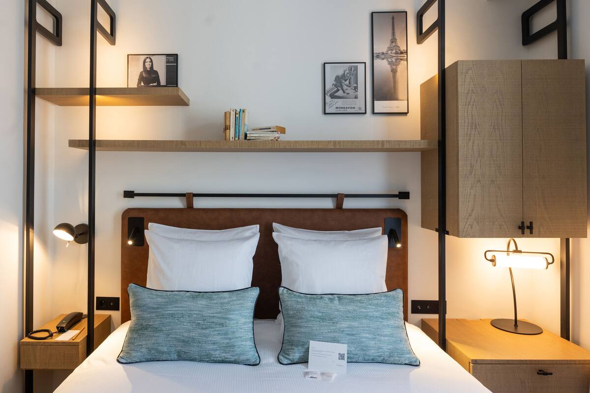 A hotel room with a bed, bedside tables, a nightstand, a lamp, a shelf with books and a framed picture, and a cabinet.