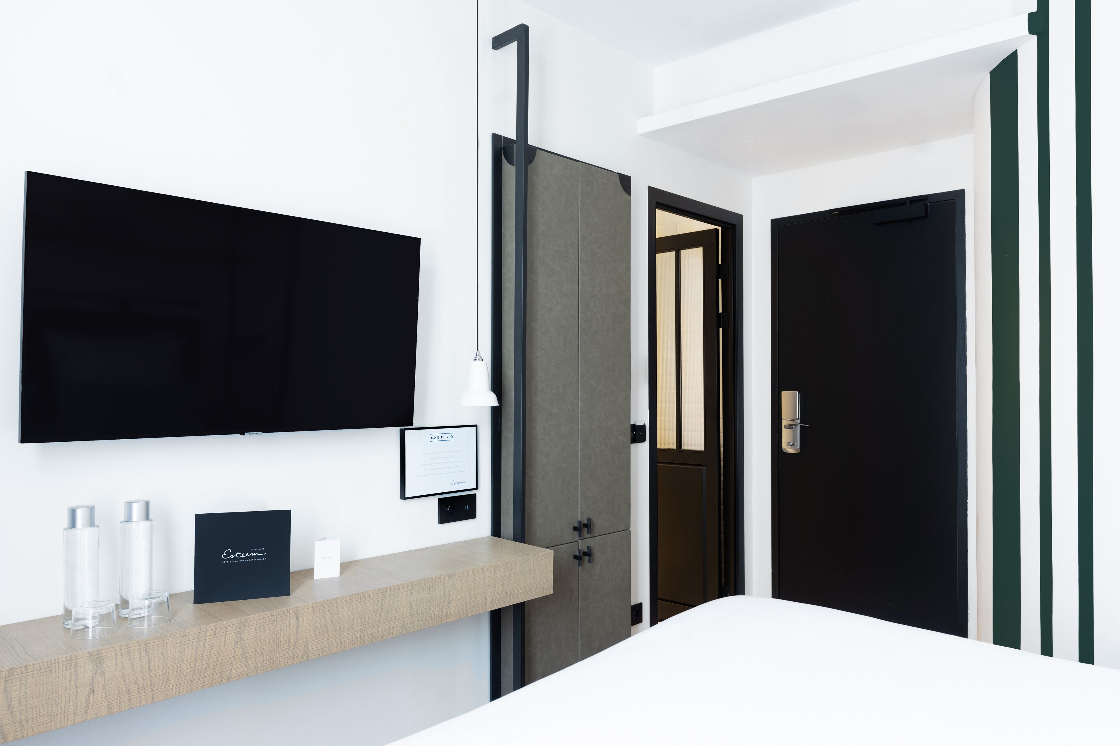 A modern hotel room with a mounted TV, a wooden shelf with toiletries, and a dark door.
