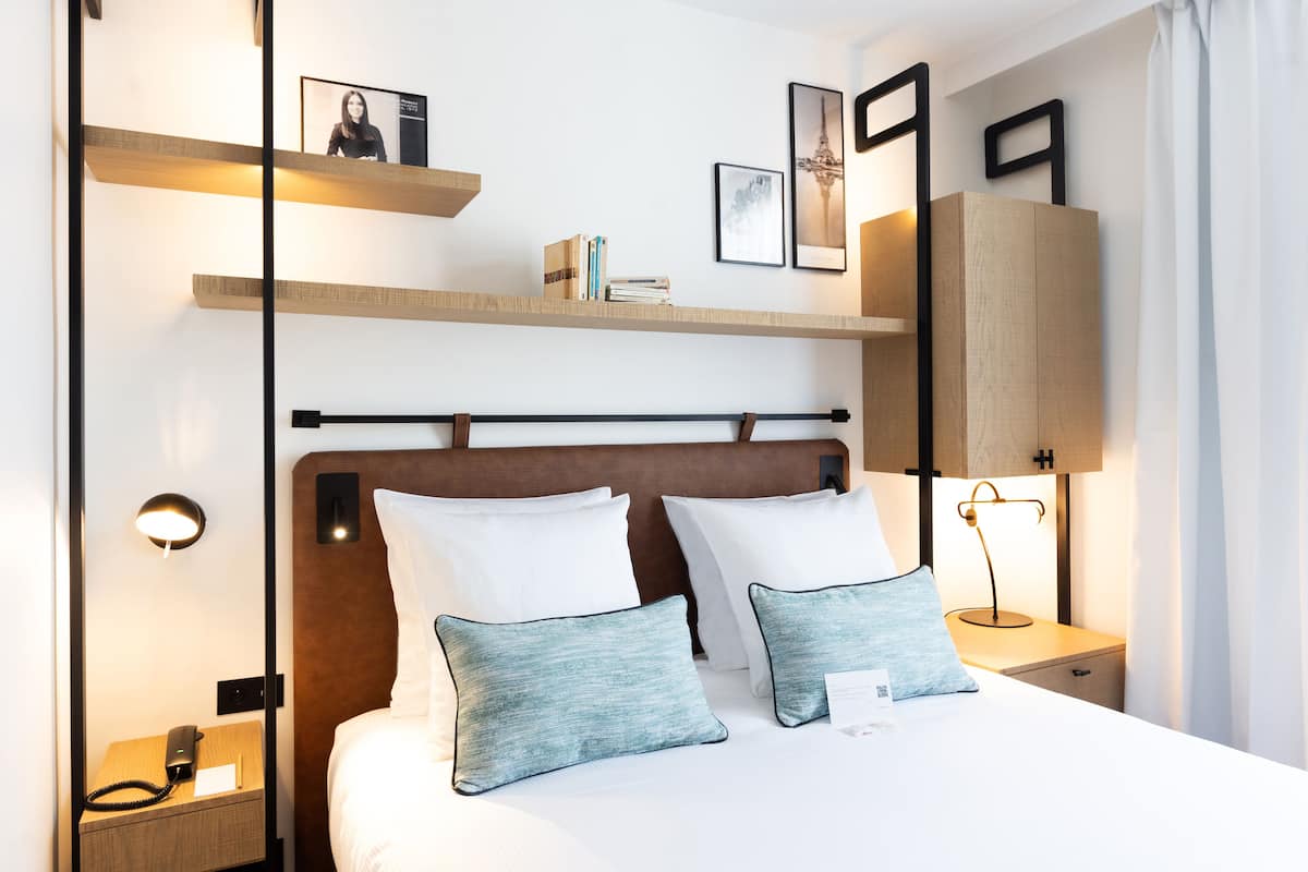 A modern hotel room with a wooden headboard, two white pillows, a bedside table with a lamp, and a shelf above the bed with books and a framed picture.