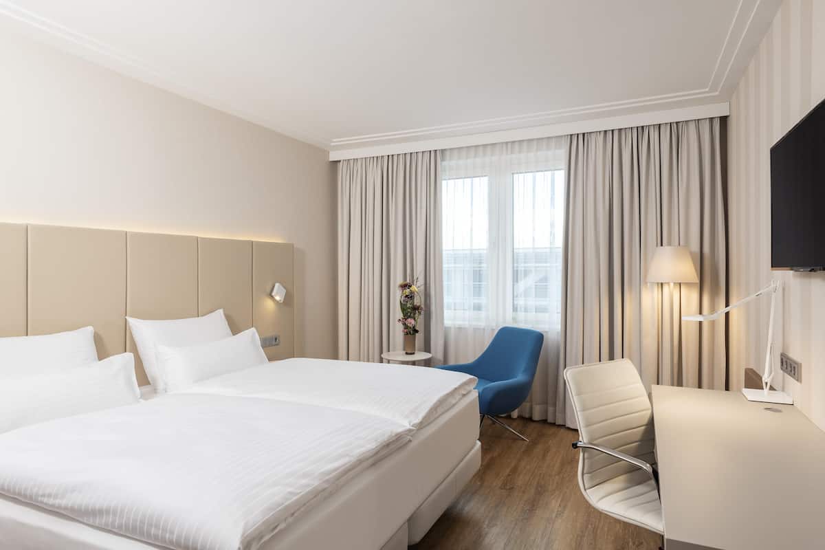 Une chambre d’hôtel avec un grand lit, un bureau, une chaise, une télévision et une fenêtre avec des rideaux.