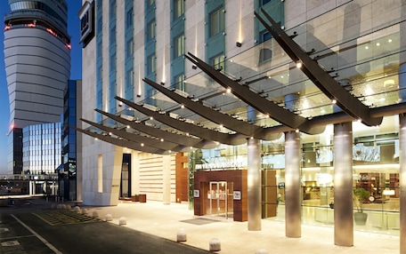 A modern hotel building with a prominent 'NH' sign, large glass windows, and a structured entrance area.