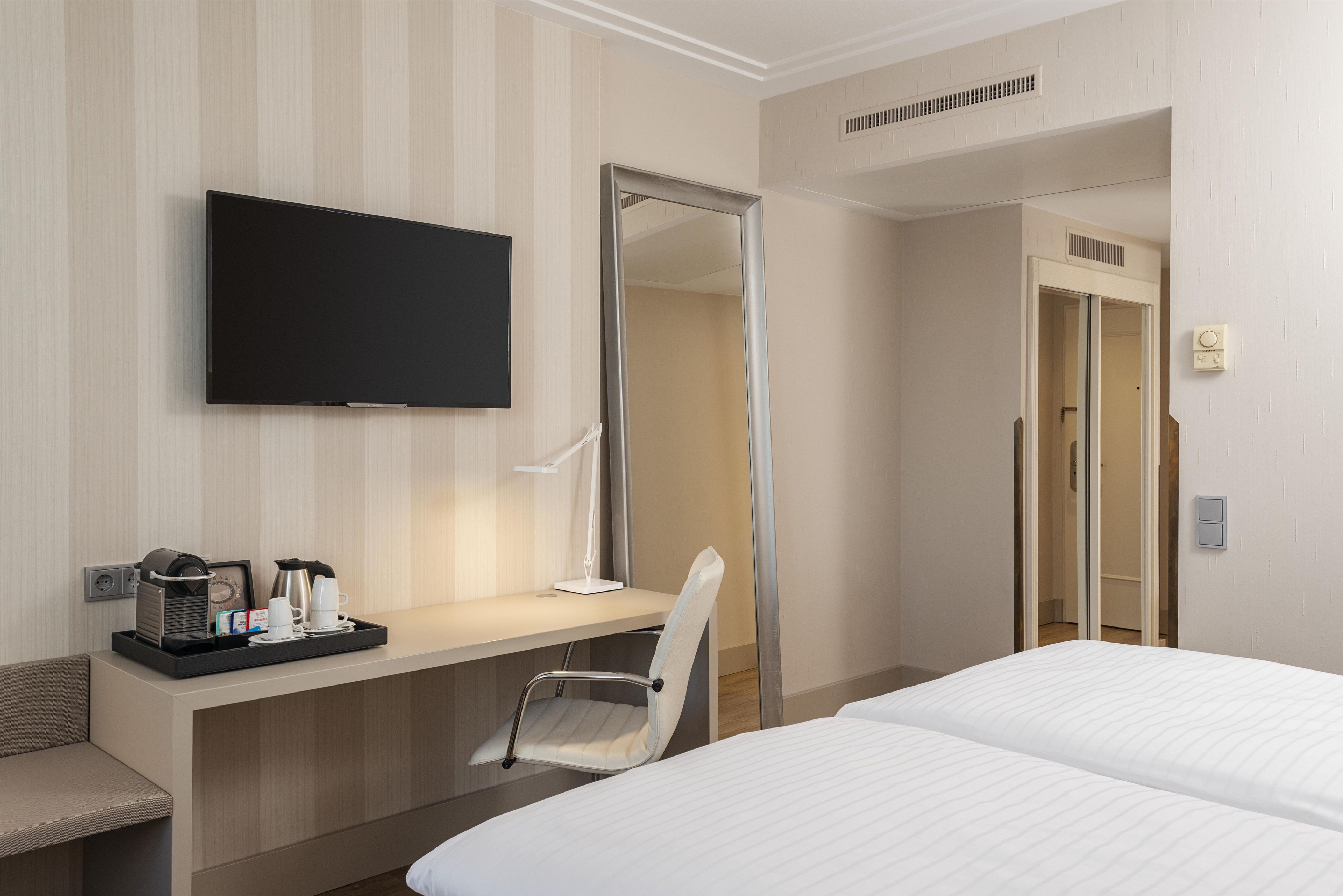 A hotel room with a bed, a desk with a coffee maker and cups, a TV, and a mirror.