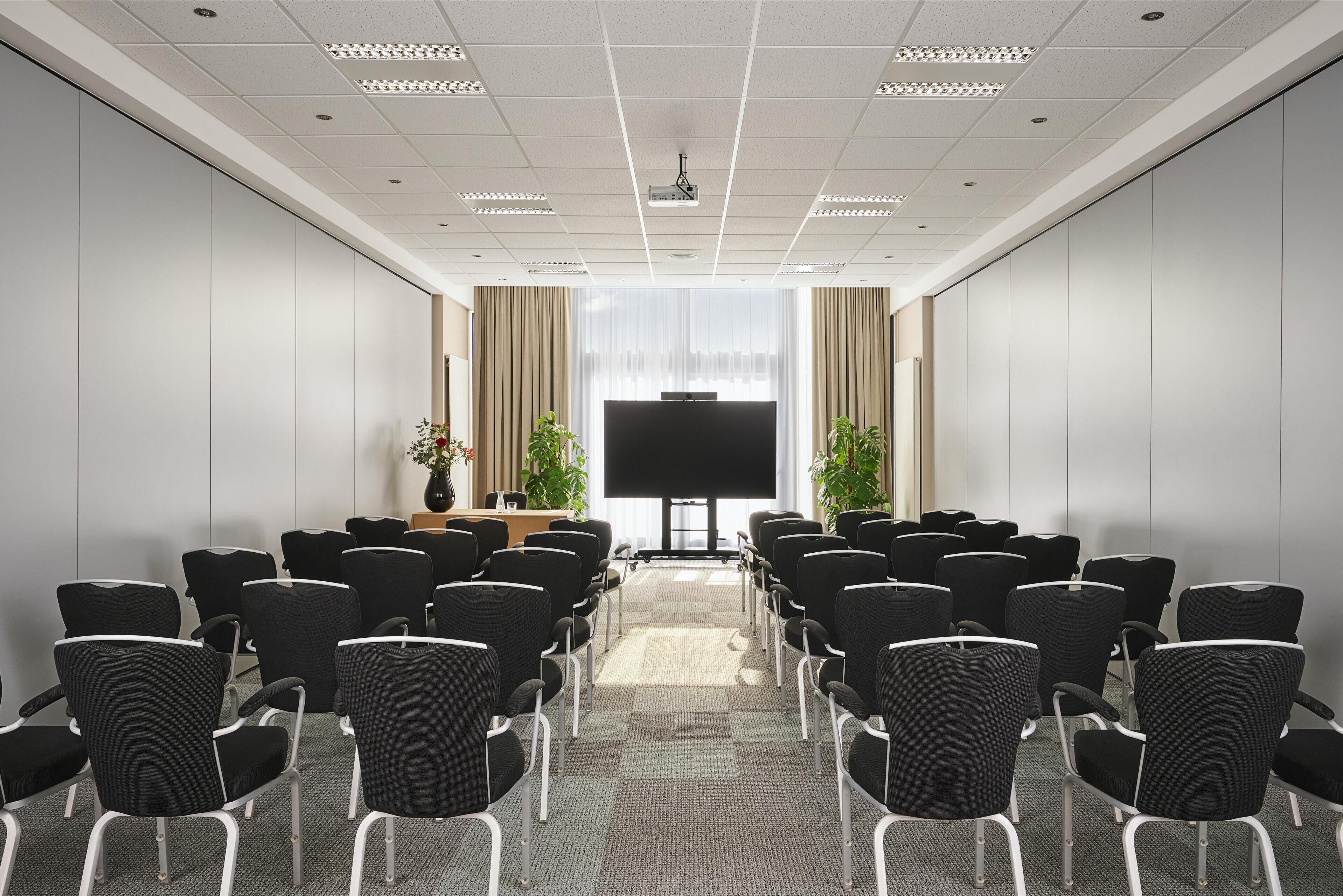 Un salón de conferencias con filas de sillas negras, un televisor de pantalla plana grande y un atril al frente.