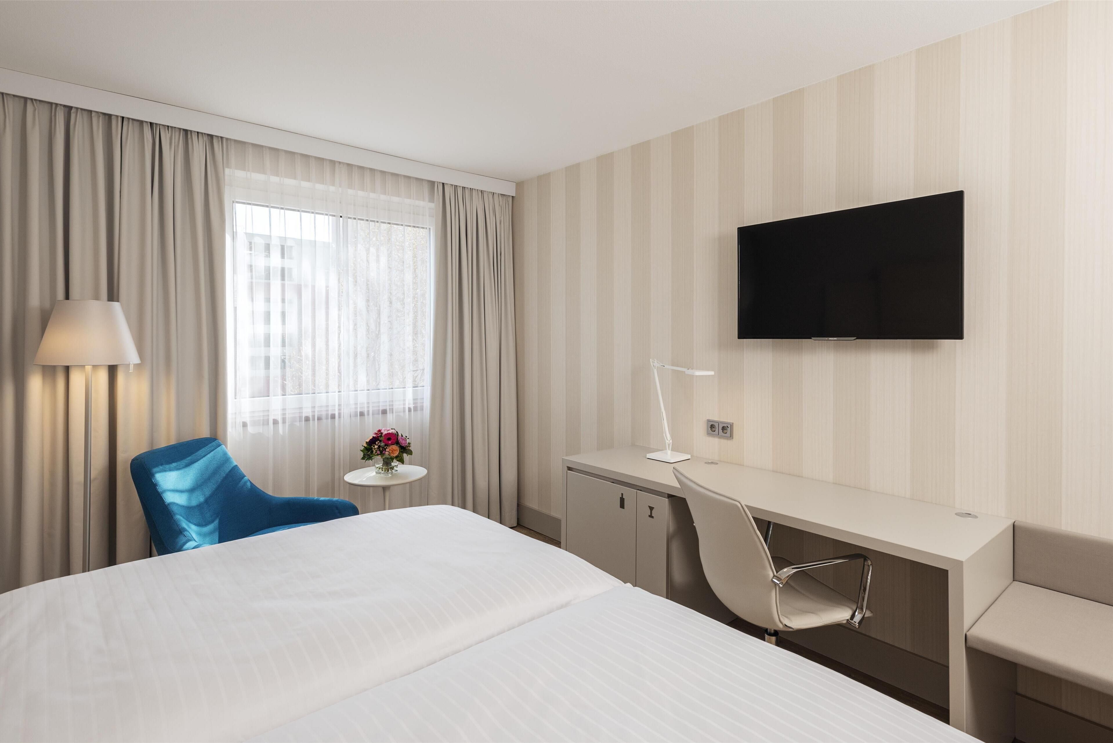 Ein Hotelzimmer mit einem großen Bett, einem Schreibtisch, einem Sessel, einem Fernseher und einem Fenster mit Vorhängen.