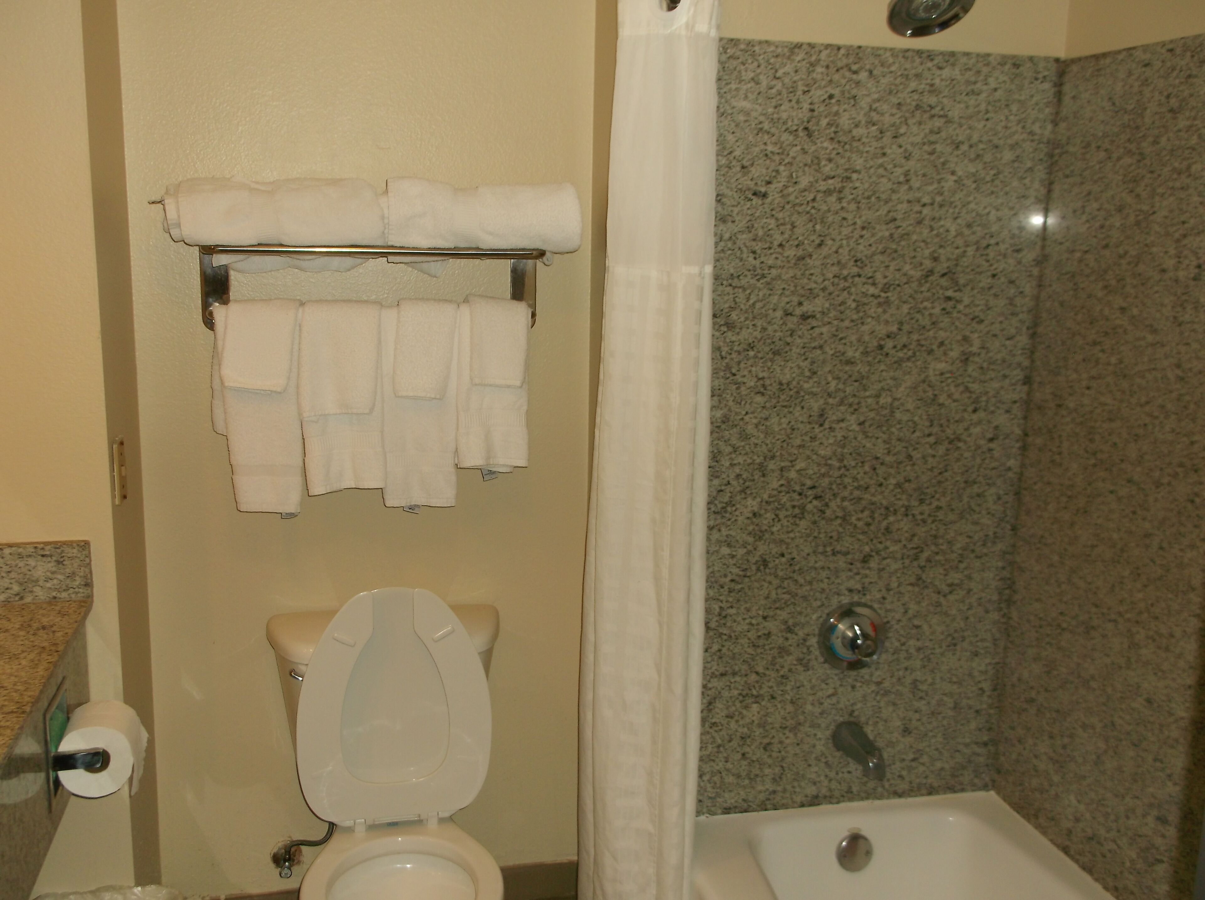 Un baño con inodoro, toallero con varias toallas, cortina de ducha y pared de ducha con azulejos de granito.