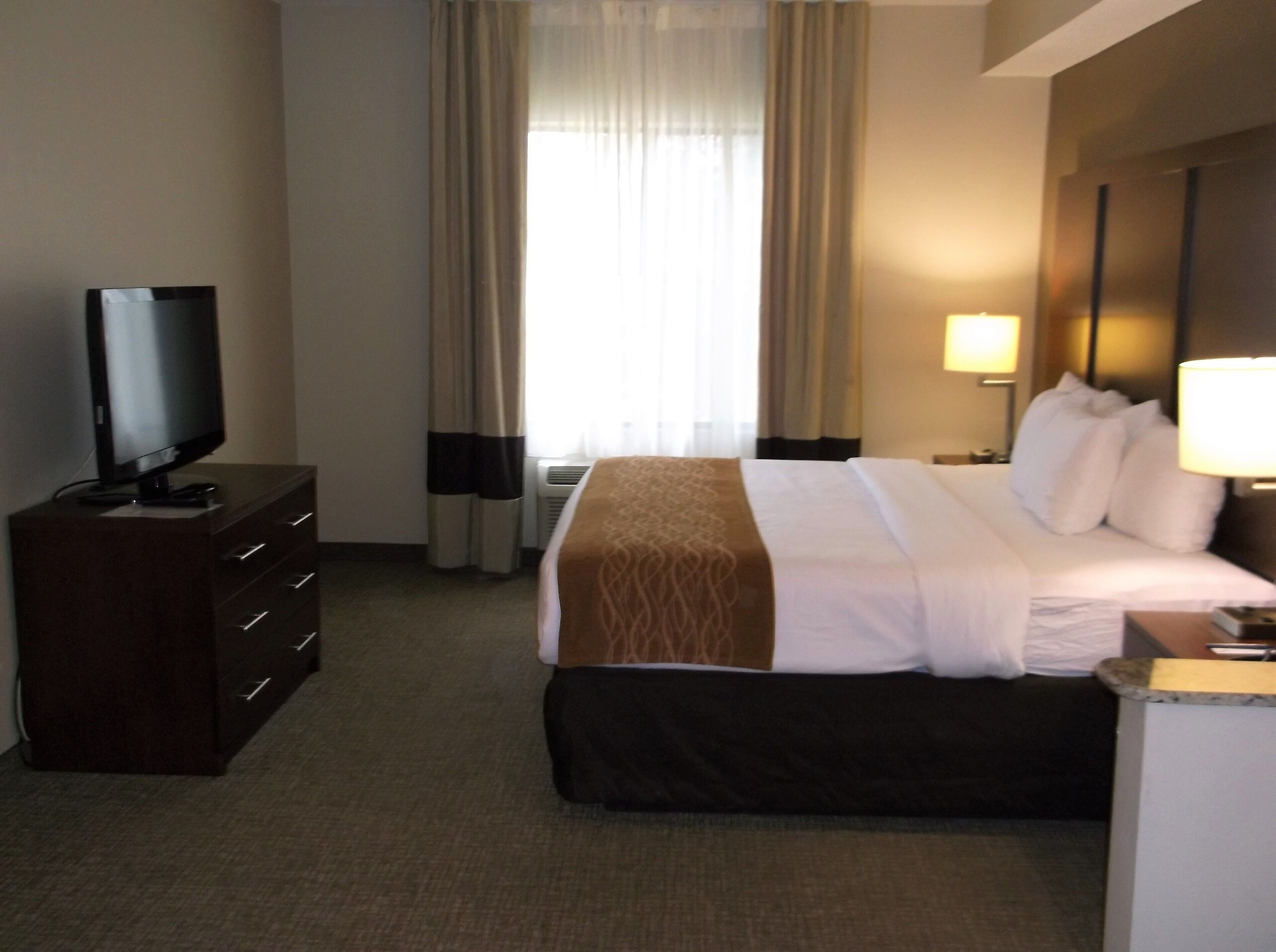 Una habitación de hotel con una cama, un televisor sobre una cómoda, una lámpara y una ventana con cortinas.