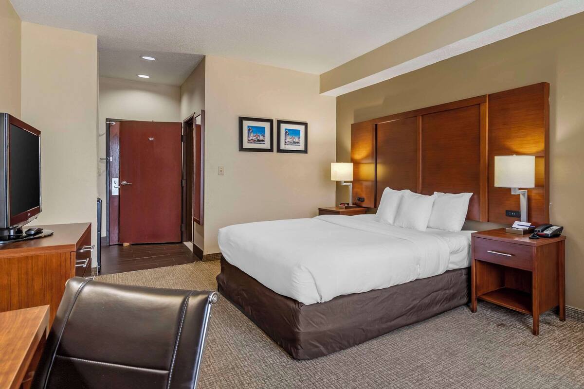 A hotel room with a large bed, a desk with a computer, a TV, a chair, and a door leading to a bathroom.