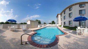 Piscina rectangular con borde de ladrillo, rodeada de sillas reclinables blancas y sombrillas azules.